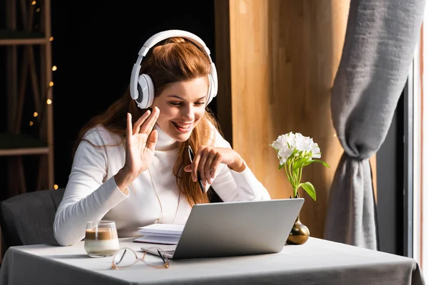 Beautiful happy woman in headphones waving and having video chat on laptop in cafe — Stock Photo
