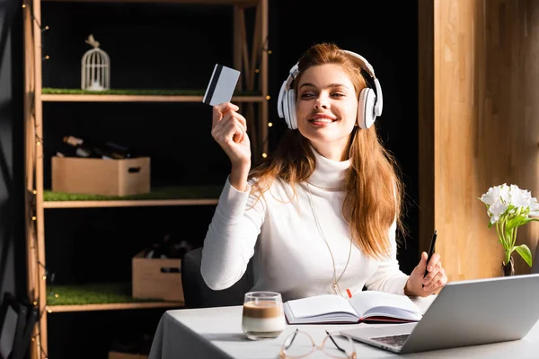 Menina ruiva sorridente em fones de ouvido pagando com cartão de crédito no café com laptop, bloco de notas e café — Fotografia de Stock