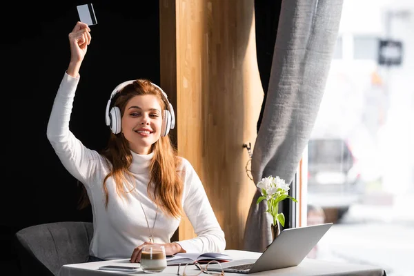 Mulher ruiva bonita em fones de ouvido pagando com cartão de crédito no café com laptop, bloco de notas e café — Fotografia de Stock