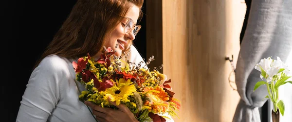 Atractiva pelirroja feliz con ramo de flores de otoño cerca de la ventana - foto de stock