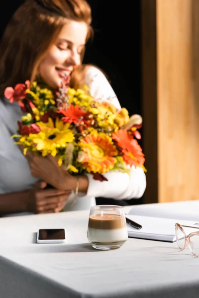 Селективное внимание счастливой женщины с букетом осенних цветов в кафе с кофейной чашкой, смартфоном и ноутбуком — стоковое фото
