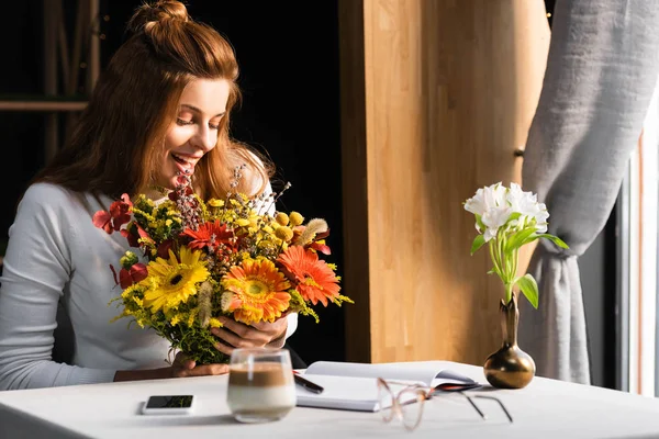 Aufgeregte rothaarige Frau mit Herbstblumenstrauß im Café mit Smartphone und Notizbuch — Stockfoto