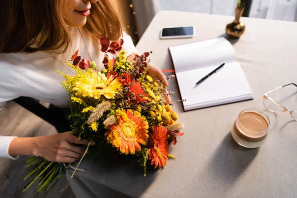 Обрезанный вид рыжеволосой женщины с букетом осенних цветов в кафе с кофе, смартфоном и ноутбуком — стоковое фото