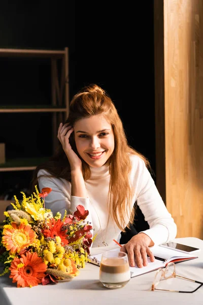 Jolie femme avec bouquet de fleurs d'automne dans un café avec smartphone, tasse de café et ordinateur portable — Photo de stock