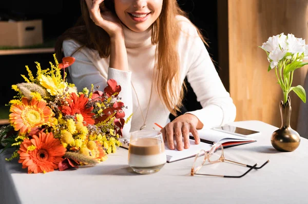 Vista ritagliata di donna con mazzo di fiori autunnali in caffè con smartphone e notebook — Foto stock