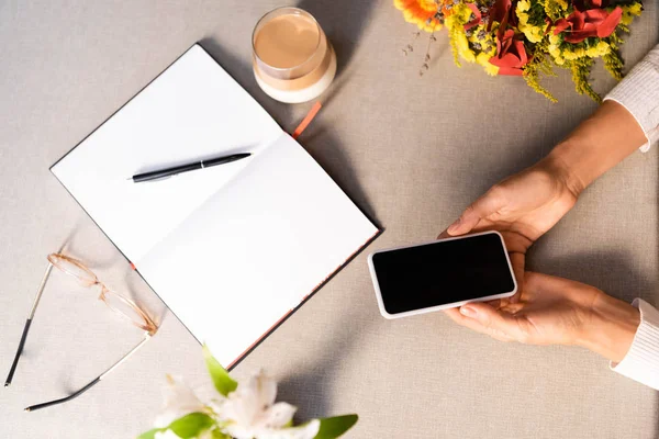 Vista cortada da mulher usando smartphone no café com bloco de notas e vidro de café — Fotografia de Stock