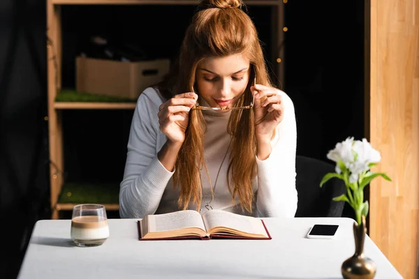 Attractive redhead woman with smartphone and glass of coffee reading book in cafe — Stock Photo
