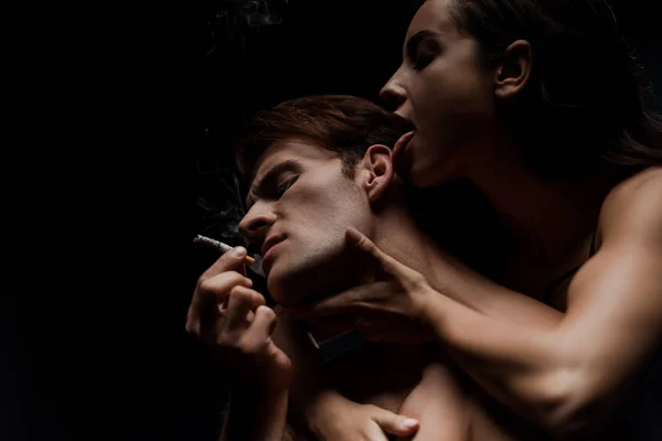 Apasionada mujer abrazando hombre fumar cigarrillo, aislado en negro - foto de stock