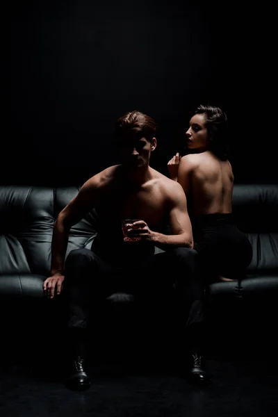 Femme sexy et homme avec un verre de whisky posant sur le canapé dans la chambre noire — Photo de stock