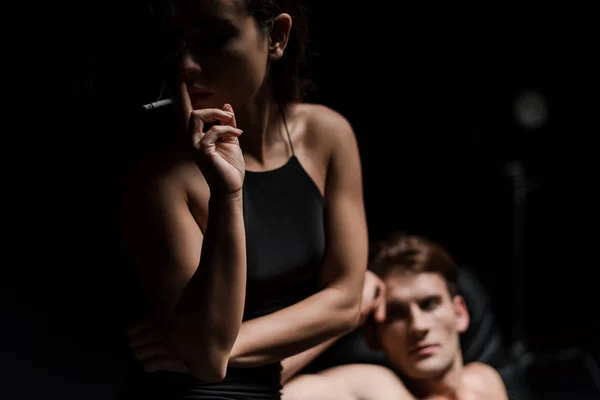 Seductive young couple sitting on sofa in dark room, while sexy woman smoking cigarette — Stock Photo