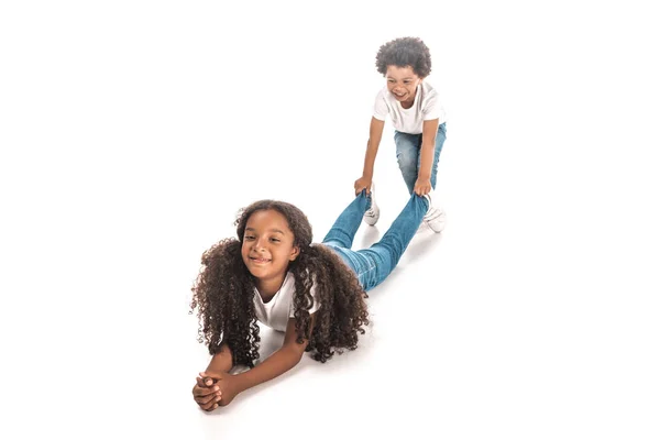 Joyeux garçon afro-américain traînant sœur par jambes sur fond blanc — Photo de stock