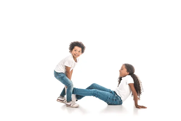 Cheerful african american boy dragging sister by leg on white background — Stock Photo