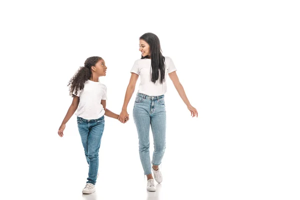 Feliz afro-americana madre e hija cogidas de la mano y mirándose mientras caminan sobre fondo blanco - foto de stock