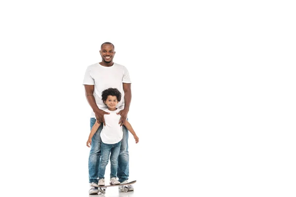 Feliz afro-americano homem de apoio alegre filho de pé no penny board no fundo branco — Fotografia de Stock