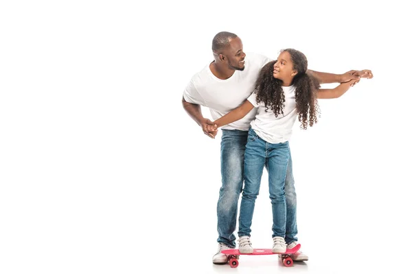 Cheerful african american father supporting adorable daughter skateboarding on white background — Stock Photo