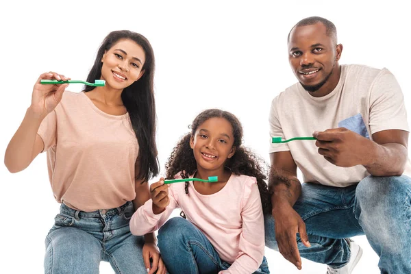 Happy african american parents with daughter holding toothbrushes and smiling at camera isolated on white — Stock Photo