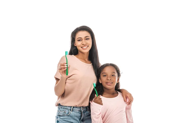 Alegre afroamericana madre e hija sosteniendo cepillos de dientes y sonriendo a cámara aislada en blanco - foto de stock