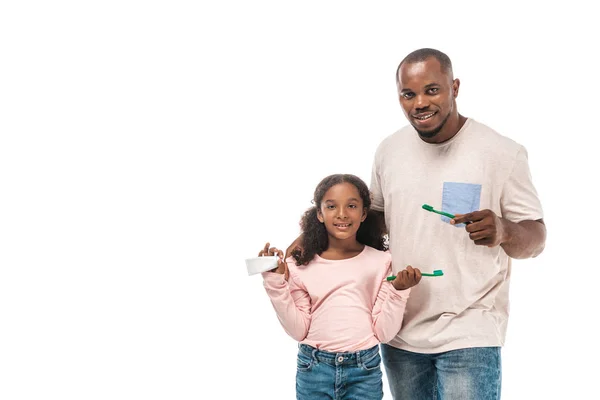 Fröhliches afrikanisch-amerikanisches Kind, das Zahnbürste und Zahnpasta in der Hand hält, während es in der Nähe des Vaters steht und die Zahnbürste isoliert auf weiß hält — Stockfoto