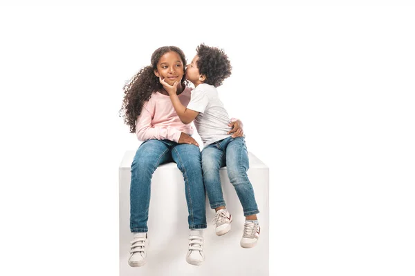 Cute african american boy kissing adorable sister while sitting on white cube together isolated on white — Stock Photo