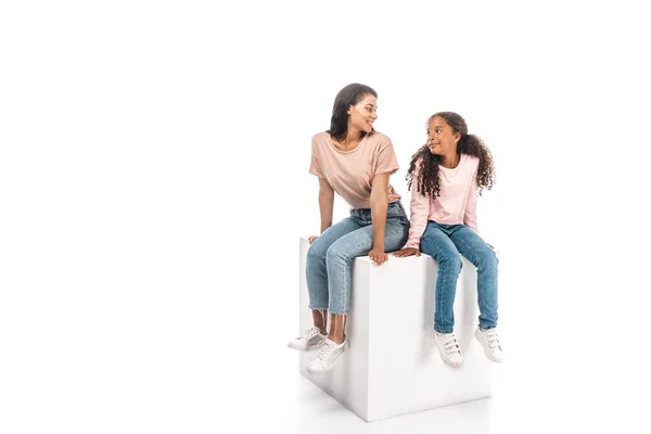Sonrientes afroamericanos madre e hija mirándose mientras están sentadas sobre un cubo blanco sobre fondo blanco - foto de stock