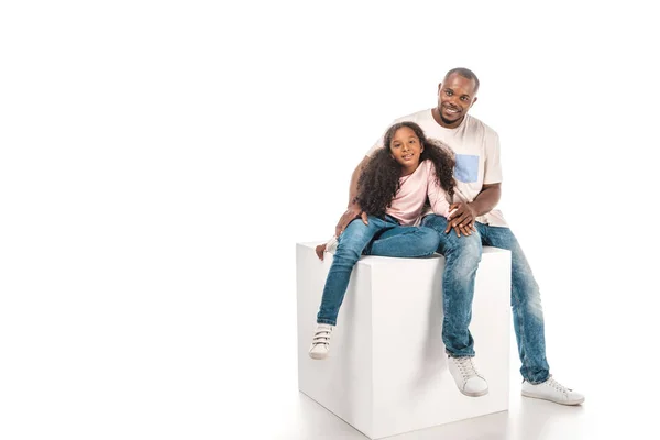Sonrientes afroamericanos padre e hija mirando a la cámara mientras están sentados en cubo juntos sobre fondo blanco - foto de stock