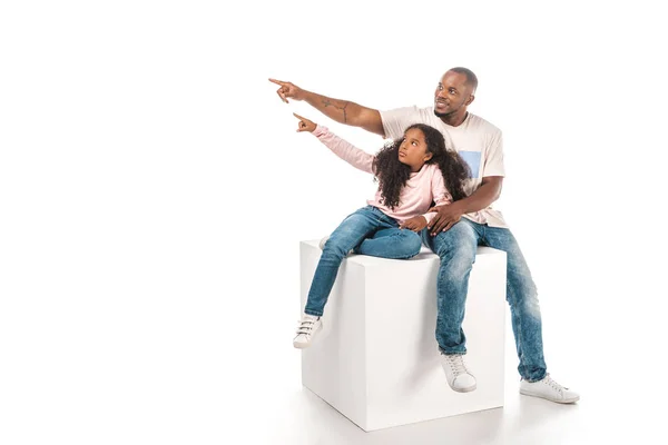 Sonrientes afroamericanos padre e hija mirando hacia otro lado y señalando con el dedo mientras están sentados en cubo juntos sobre fondo blanco - foto de stock