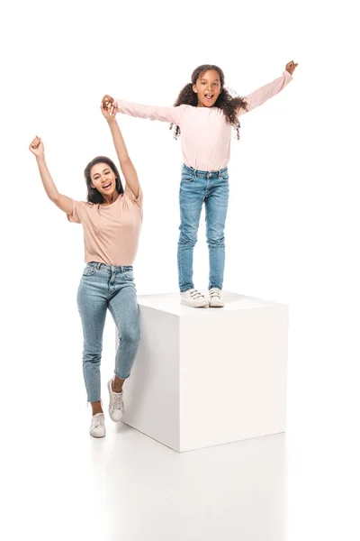 Fröhliche afrikanisch-amerikanische Frau zeigt Siegergeste, während sie die Hand der glücklichen Tochter hält, die auf einem Podest auf weißem Hintergrund steht — Stockfoto