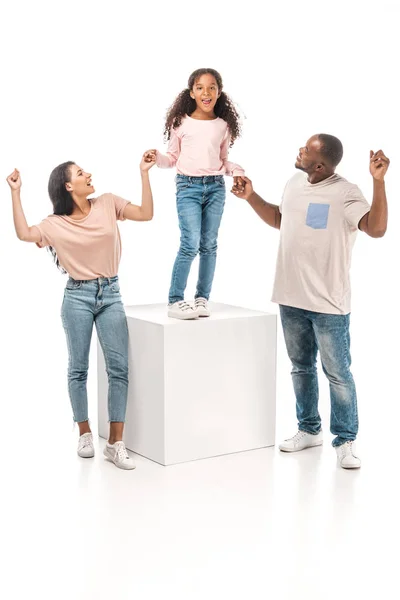 Glückliche afrikanisch-amerikanische Eltern halten Hände einer fröhlichen Tochter, die auf einem weißen Würfel auf weißem Hintergrund steht — Stockfoto