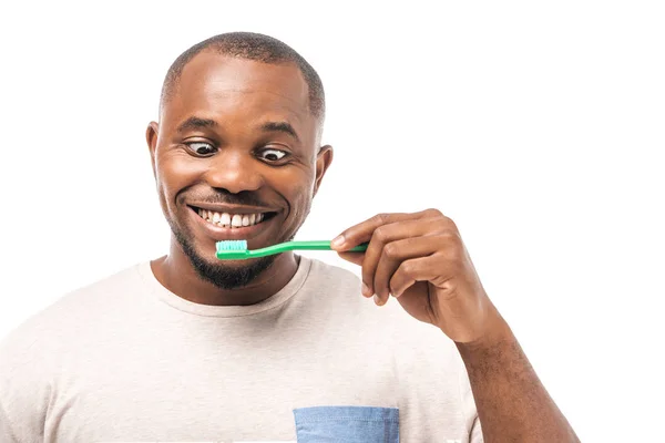 Cheerful african american man looking at toothbrush while brushing teeth isolated on white — Stock Photo