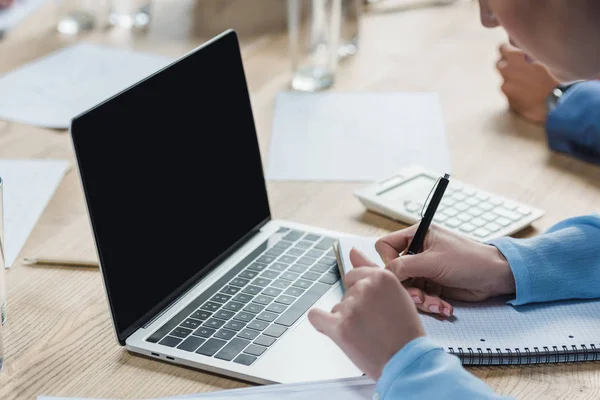 Vista recortada de la mujer de negocios sentada cerca de la computadora portátil y witing en el cuaderno durante la reunión de negocios - foto de stock