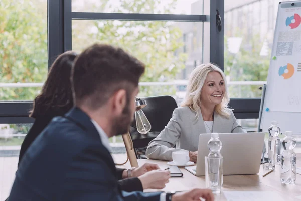 Избирательный фокус привлекательной деловой женщины, улыбающейся во время деловой встречи с молодыми коллегами — стоковое фото