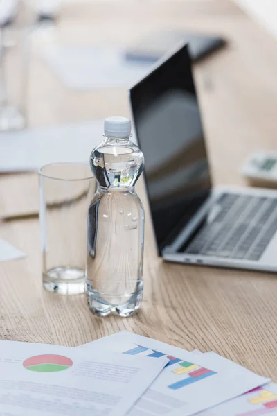 Fuoco selettivo di bottiglia e vetro d'acqua vicino a computer portatile e carte con infografica su tavolo di legno in sala riunioni — Foto stock