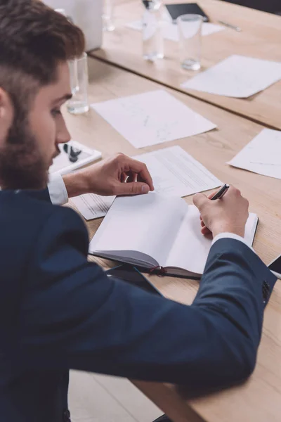 Junger Geschäftsmann schreibt in Notizbuch, während er am Schreibtisch im Besprechungsraum sitzt — Stockfoto