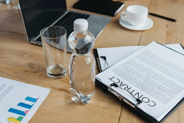 Klemmbrett mit Vertrag, Flasche Wasser und Glas, Laptop und Kaffeetasse auf Holztisch — Stockfoto
