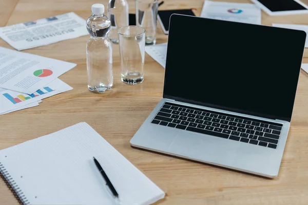 Laptop with blank screen, empty notebook, papers with infographics, bottle and glass of water on wooden table in meeting room — Stock Photo