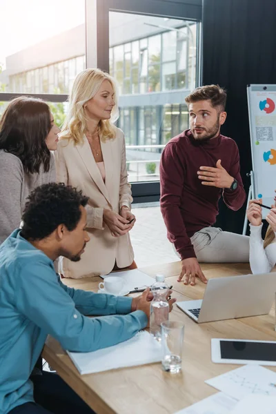 Jóvenes empresarios hablando con una mujer de negocios madura mientras que un hombre de negocios afroamericano sentado en el escritorio en la reunión de negocios - foto de stock