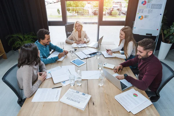 Empresários multiculturais positivos sentados em uma mesa de madeira perto de dispositivos digitais, documentos e copos com água na sala de reuniões — Fotografia de Stock