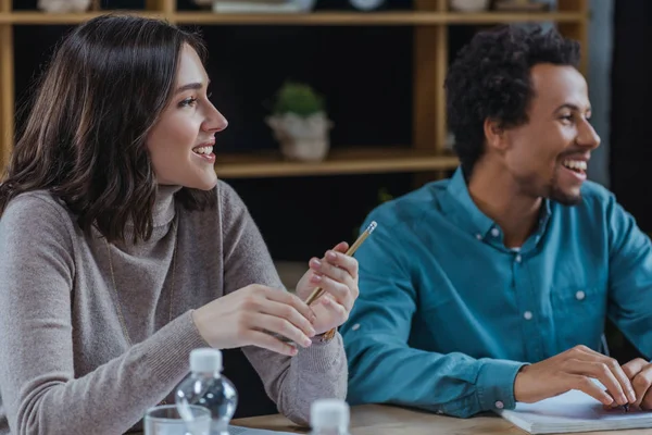 Young businesswoman holding pen while sitting near african american colleague — Stock Photo