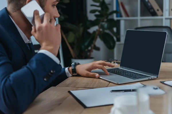 Jovem empresário sentado perto do laptop com tela em branco e falando no smartphone — Fotografia de Stock