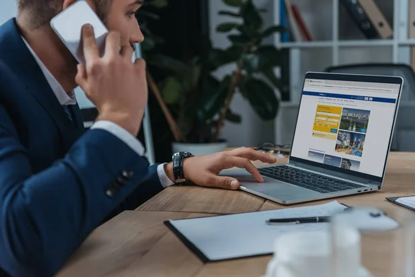 KYIV, UKRAINE - OCTOBER 1, 2019: Young businessman sitting near laptop with Booking.com website on screen and talking on smartphone. — Stock Photo