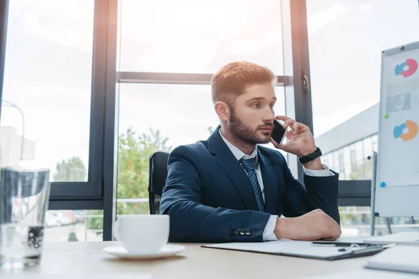 Joven hombre de negocios hablando en el teléfono inteligente mientras está sentado en el escritorio en la sala de reuniones - foto de stock