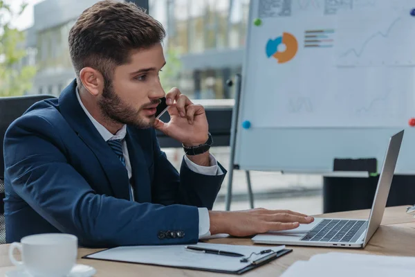 Empresário atencioso falando no smartphone enquanto usa laptop no escritório — Fotografia de Stock