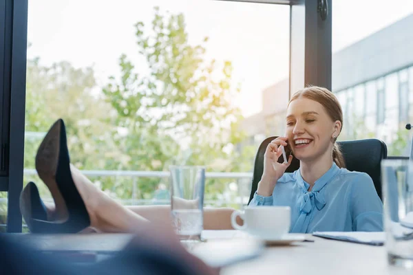 Cheerful businesswoman sitting with legs on desk and talking on smartphone — Stock Photo