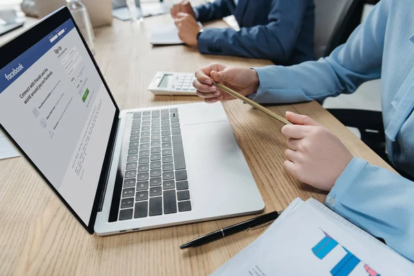 KYIV, UCRANIA - 1 de octubre de 2019: Vista recortada del hombre de negocios sentado en la sala de reuniones cerca de la computadora portátil con el sitio web de Facebook en la pantalla . - foto de stock