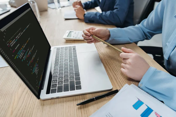 Vista recortada del programador sentado en la sala de reuniones cerca del ordenador portátil con código de programación en la pantalla - foto de stock