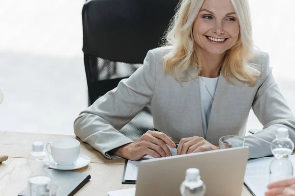 Attraktive, reife Geschäftsfrau lächelt, während sie im Besprechungsraum am Anzeigentisch sitzt — Stockfoto