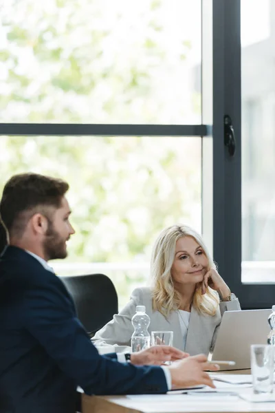 Mujer de negocios positiva y atenta escuchando a un joven colega durante la reunión de negocios - foto de stock