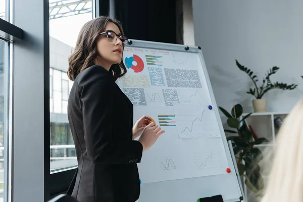 Joven mujer de negocios de pie cerca de flipchart con infografías durante la reunión de negocios - foto de stock