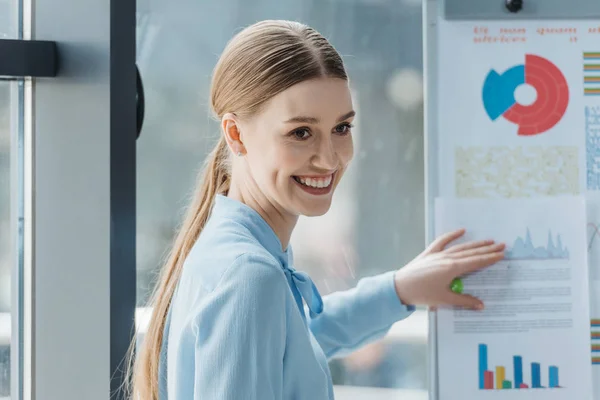 Mulher de negócios sorridente de pé perto flipchart com infográficos durante reunião de negócios — Fotografia de Stock