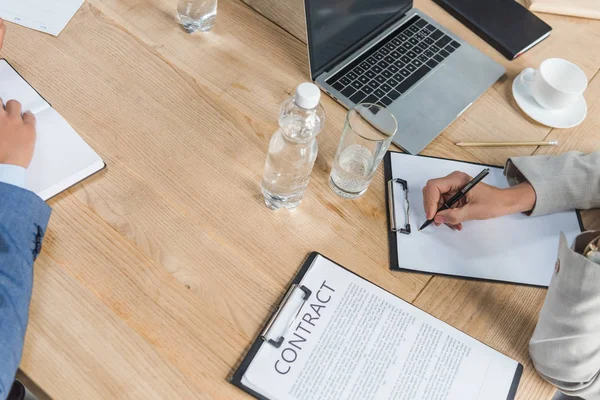 Teilansicht einer Geschäftsfrau, die auf Klemmbrett neben Laptop schreibt und Vertrag auf Holztisch — Stockfoto